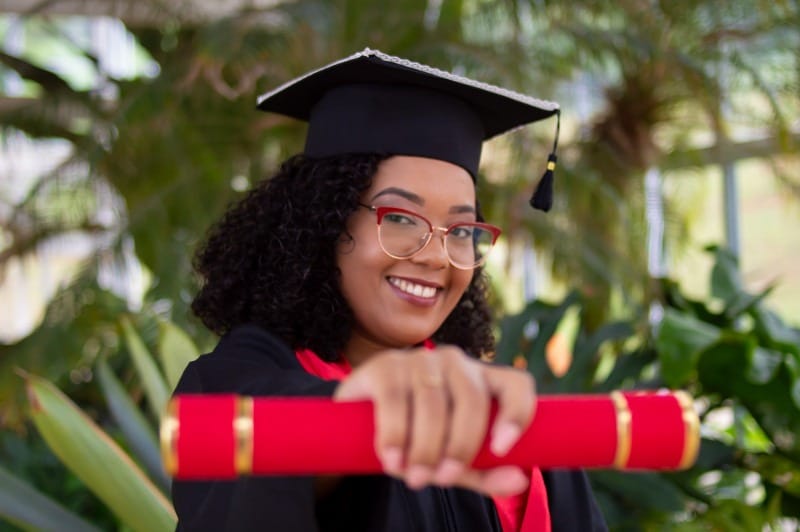 Foto de una mujer sonriendo con su diploma de graduación en la mano, vistiendo toga y birrete